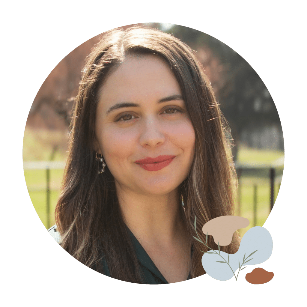 Dr. Jennifer Melo, Psychologist PsD. for trauma informed healing. A woman with long, wavy brown hair smiles at the camera. She is wearing a dark green collared shirt and subtle earrings. The background is blurred with trees and a fence.