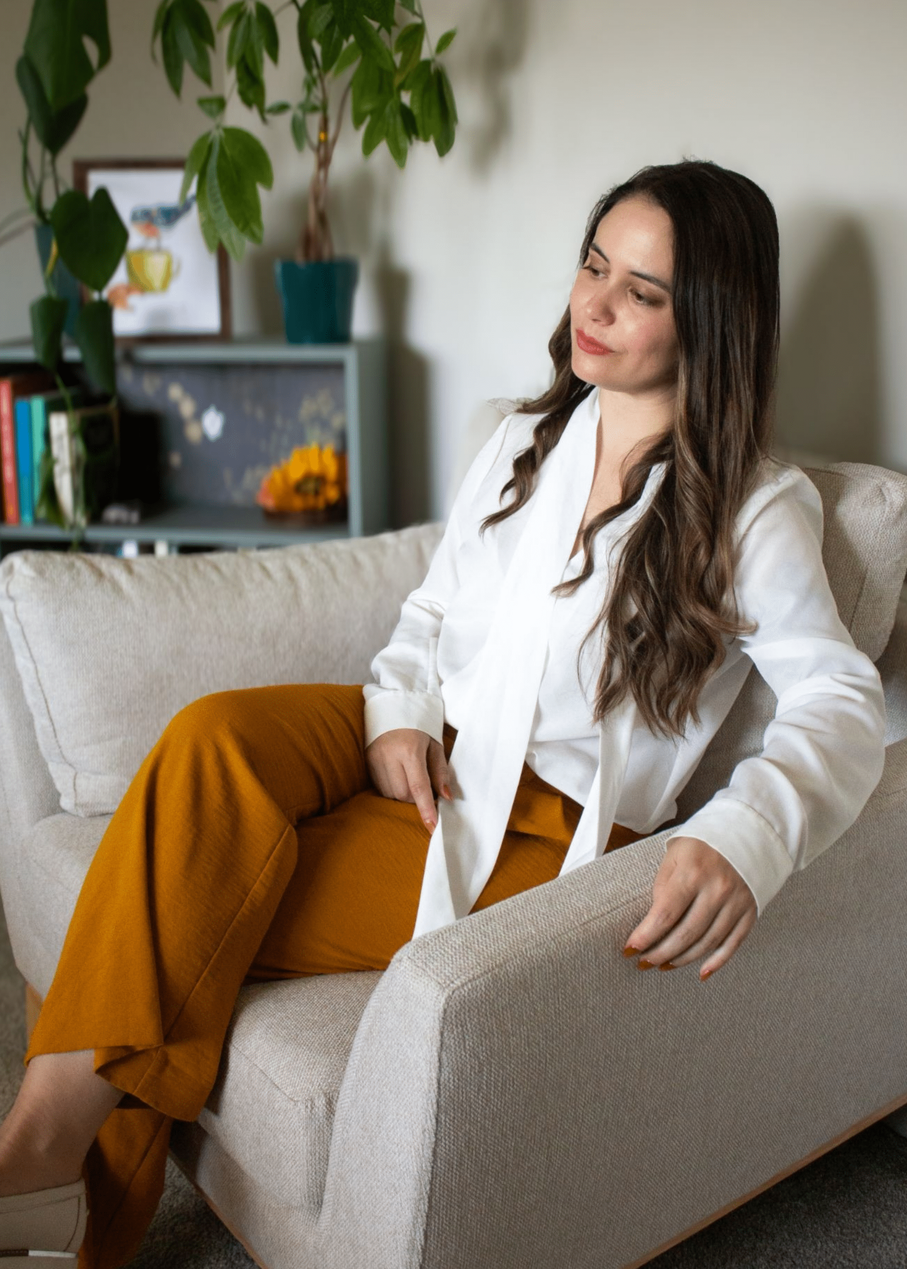 Dr. Jennifer Melo wearing a white blouse and mustard-colored pants sits in a beige armchair, with a bookshelf and plants in the background. Psychologist in DMV area