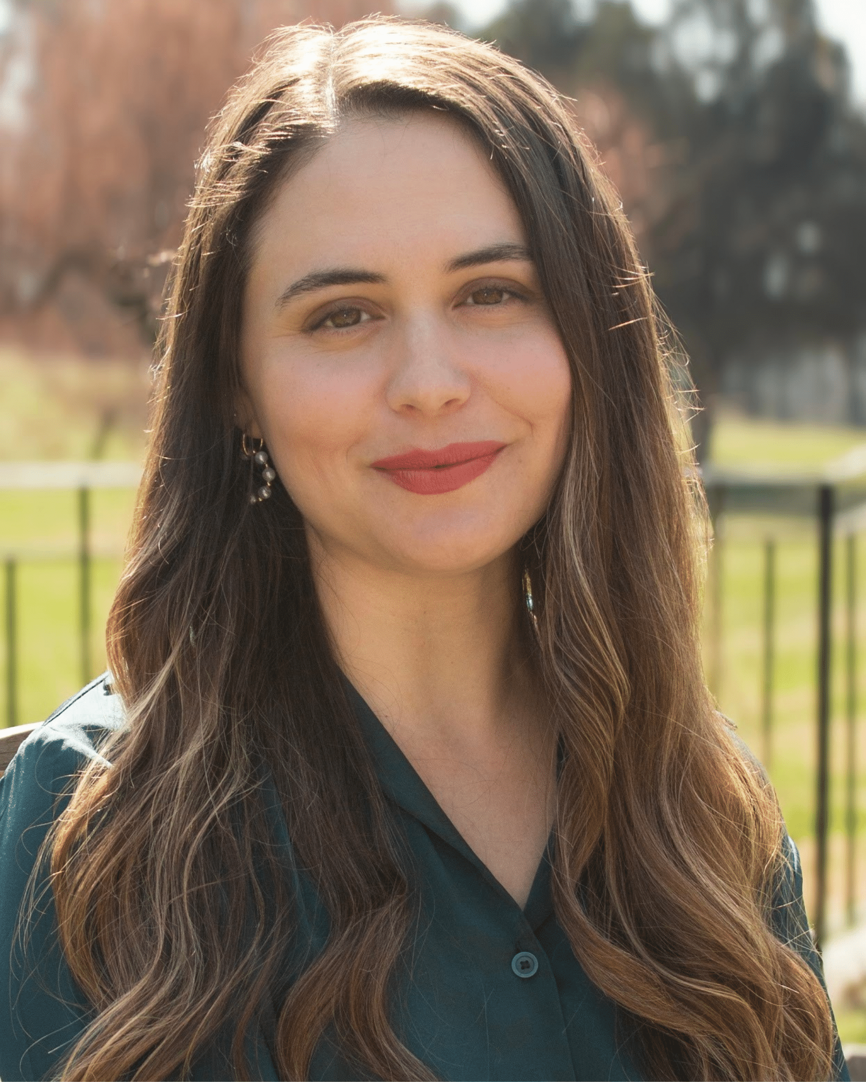 Dr. Jennifer Melo, Psychologist PsD. for trauma informed healing. A woman with long, wavy brown hair smiles at the camera. She is wearing a dark green collared shirt and subtle earrings. The background is blurred with trees and a fence.